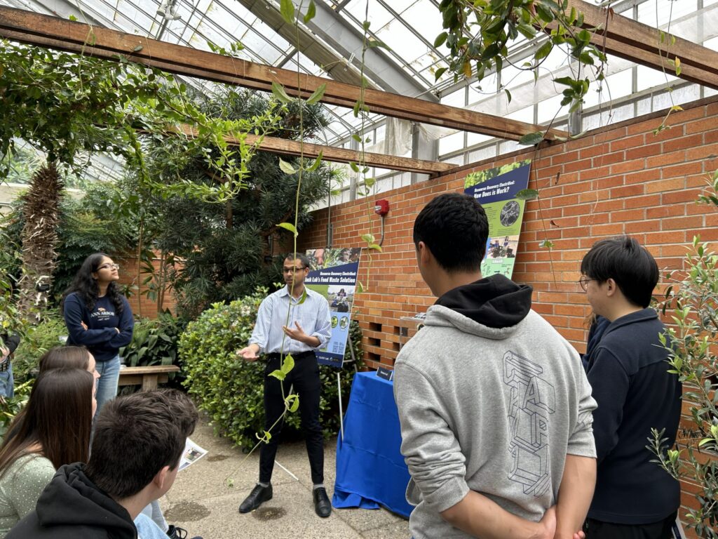 A student presents research to a small group in a greenhouse surrounded by plants and posters.